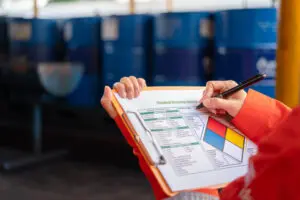 A safety officer is checking on the hazardous material checklist form with chemical storage area at the factory as background. Industrial safety working scene, selective focus.